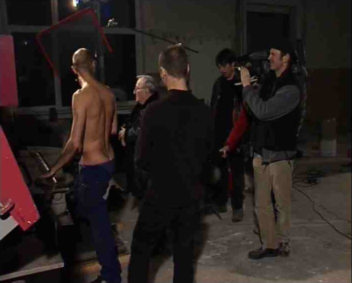 Gad Beck inszeniert eine erotische Begegnung aus seiner Zeit als Zwangsarbeiter. Es ist ein Mann mit Glatze und nacktem Oberkörper zu sehen, Gad Beck gibt ihm Regieanweisungen. Drumherum steht ein Filmteam mit Kamera und Mikrofonangel